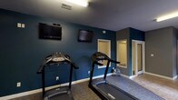 The Reserve's fitness center is a thoughtfully designed, single-floor facility organized across multiple interconnected rooms, providing residents with a comprehensive space for a full-body workout. The main cardio area features multiple treadmills positioned beneath wall-mounted flat-screen televisions, making it easy to stay entertained during endurance training. Bold teal accent walls contrast with neutral gray tones throughout, giving the space a modern, energetic atmosphere complemented by durable carpet flooring suited for athletic use.

A dedicated room houses an elliptical cross-trainer alongside a wall-mounted accessory rack, with natural light filtering in through window blinds to create a bright, welcoming environment. An adjacent strength training room provides open floor space with a weight bench and free weights, offering ample room for resistance and functional fitness exercises. This area also benefits from natural light through a window and features the same consistent gray color palette and carpet flooring found throughout the facility.

A tiled hallway connects the various workout areas, providing smooth transitions between zones and leading to additional amenities visible along the corridor. The overall layout is practical and efficient, ensuring users can move seamlessly between cardio, strength, and open training areas within one cohesive fitness facility.