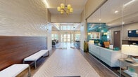 This generously sized amenity facility in Lynnfield, MA offers a thoughtfully designed, single-floor layout encompassing a diverse range of communal spaces. The entrance greets visitors with soaring double-height ceilings, an abundance of natural light pouring through oversized windows, and elegant herringbone-patterned tile flooring that flows throughout the main corridors and common areas.

The living room is a standout gathering space, featuring a coffered ceiling, a central fireplace flanked by built-in shelving, large windows with garden views, and comfortable seating arranged around a statement area rug. Adjacent to it, the modern kitchen boasts white cabinetry, a textured tile backsplash, stainless steel appliances, and a granite-topped island with bar seating illuminated by pendant lighting. A dedicated dining room offers a sophisticated setting with a glass-top table, emerald-green accent chairs, and curated artwork.

The fitness center is expansive and well-appointed, featuring rows of cardio equipment including treadmills and stationary bikes, strength training machines, floor-to-ceiling mirrors, and vibrant patterned carpet. A glass-walled corridor connects the gym to the main hall, where package concierge lockers provide added convenience. The facility also offers direct access to an outdoor pool and lounge deck, completing this comprehensive amenity offering.