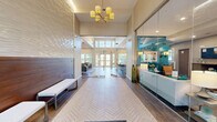 This generously sized amenity facility in Lynnfield, MA offers a thoughtfully designed, single-floor layout encompassing a diverse range of communal spaces. The entrance greets visitors with soaring double-height ceilings, an abundance of natural light pouring through oversized windows, and elegant herringbone-patterned tile flooring that flows throughout the main corridors and common areas.

The living room is a standout gathering space, featuring a coffered ceiling, a central fireplace flanked by built-in shelving, large windows with garden views, and comfortable seating arranged around a statement area rug. Adjacent to it, the modern kitchen boasts white cabinetry, a textured tile backsplash, stainless steel appliances, and a granite-topped island with bar seating illuminated by pendant lighting. A dedicated dining room offers a sophisticated setting with a glass-top table, emerald-green accent chairs, and curated artwork.

The fitness center is expansive and well-appointed, featuring rows of cardio equipment including treadmills and stationary bikes, strength training machines, floor-to-ceiling mirrors, and vibrant patterned carpet. A glass-walled corridor connects the gym to the main hall, where package concierge lockers provide added convenience. The facility also offers direct access to an outdoor pool and lounge deck, completing this comprehensive amenity offering.