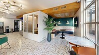 Located at 2024 Durant Ave in Berkeley, CA, this thoughtfully designed communal lounge space offers a compelling blend of modern aesthetics and practical amenities. The space is unified by striking terrazzo flooring throughout, which adds texture and visual depth while complementing the eclectic, design-forward interior.

The main lounge area features bold navy and teal accent walls, a wall-mounted flat-screen TV set within a warm wood-framed niche, and sculptural black-and-gold sputnik chandeliers that provide both ambient and accent lighting. An Aquafina beverage refrigerator offers convenient refreshment access, while a dedicated kitchenette area showcases sage green cabinetry with brushed gold hardware, open shelving adorned with blue subway tile, and a sink with a gold faucet — all contributing to an elevated, boutique-style finish.

A self-contained privacy pod with glass panels provides a quiet enclave within the open layout, ideal for focused work or calls. A cushioned built-in banquette with decorative patterned upholstery and warm wall sconces creates a cozy lounge nook, complemented by marble-topped tables and lush potted tropical plants. Floor-to-ceiling windows and glass doors flood the interior with natural light and offer views of an outdoor terrace area, seamlessly connecting the interior with the exterior environment.