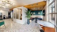 Located at 2024 Durant Ave in Berkeley, CA, this thoughtfully designed communal lounge space offers a compelling blend of modern aesthetics and practical amenities. The space is unified by striking terrazzo flooring throughout, which adds texture and visual depth while complementing the eclectic, design-forward interior.

The main lounge area features bold navy and teal accent walls, a wall-mounted flat-screen TV set within a warm wood-framed niche, and sculptural black-and-gold sputnik chandeliers that provide both ambient and accent lighting. An Aquafina beverage refrigerator offers convenient refreshment access, while a dedicated kitchenette area showcases sage green cabinetry with brushed gold hardware, open shelving adorned with blue subway tile, and a sink with a gold faucet — all contributing to an elevated, boutique-style finish.

A self-contained privacy pod with glass panels provides a quiet enclave within the open layout, ideal for focused work or calls. A cushioned built-in banquette with decorative patterned upholstery and warm wall sconces creates a cozy lounge nook, complemented by marble-topped tables and lush potted tropical plants. Floor-to-ceiling windows and glass doors flood the interior with natural light and offer views of an outdoor terrace area, seamlessly connecting the interior with the exterior environment.