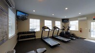 Located at 1524 Braxton Pkwy in Columbus, Ohio, this fitness center offers a well-appointed, single-floor workout space designed to meet a variety of exercise needs. The room features a clean, neutral color palette with beige walls and a carpeted floor, creating a functional yet comfortable environment for physical activity. Natural light streams in through multiple white-framed windows fitted with blinds, giving the space an open and airy feel. Recessed ceiling lighting provides consistent illumination throughout. The cardio section is equipped with multiple treadmills and an elliptical trainer, ideal for aerobic workouts. A full dumbbell rack lines one wall, offering a range of free weights for strength training. Two wall-mounted flat-screen televisions — including a U-verse connected display — are positioned for easy viewing from the cardio equipment, making workouts more engaging. The layout is thoughtfully organized to maximize floor space and allow for comfortable movement between stations. Overall, this fitness center provides a practical and accessible workout environment suitable for residents or members seeking a convenient on-site exercise facility.