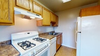 This single-floor residence offers a practical and well-organized layout that balances comfort with functionality. The kitchen is efficiently designed, featuring warm honey-toned wood cabinetry, dark granite-style countertops, and a full suite of white appliances including a gas range, dishwasher, and refrigerator. An overhead ventilation hood and recessed upper cabinets maximize storage, while durable vinyl flooring completes the space.

The living area is generously proportioned, featuring plush grey carpeting and a large window dressed with vertical blinds that admits abundant natural light and frames outdoor views. A ceiling fan provides additional comfort overhead, while a textured ceiling adds subtle architectural detail throughout the common areas.

The bedrooms are well-sized, each finished with the same soft grey carpeting and neutral-toned walls. Built-in closets with shelving provide organized storage solutions, and windows with vertical blinds allow for natural light control.

The bathroom is clean and straightforward, featuring white tile surrounding a full soaking tub and shower, white tile flooring, a single-sink vanity with wood cabinetry, a large mirror, and overhead lighting. The overall ambiance of the space is neutral and move-in ready, offering a blank canvas suitable for a variety of furnishing styles.