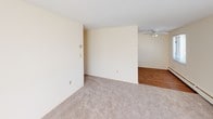 This single-floor residence offers a clean, functional layout designed for comfortable everyday living. The living area is generously sized, featuring soft wall-to-wall carpeting, neutral cream-toned walls, and multiple windows fitted with blinds that allow for ample natural light. Baseboard heating and a wall-mounted air conditioning unit ensure year-round climate control throughout the space. A ceiling fan with integrated lighting adds both comfort and practicality to the main living zone. The kitchen is outfitted with warm honey-toned wood cabinetry, light countertops, a double stainless steel sink, and hardwood-style flooring that adds a touch of warmth. It accommodates a full range of appliances, including a dishwasher and electric range. The bedroom is a comfortable retreat with plush carpeting, natural light from a window with blinds, and direct access to both a well-sized closet and a full bathroom. The walk-in closet features wire shelving on multiple sides, providing organized and practical storage. The bathroom is finished with white square tile surrounding the bathtub and shower, a large vanity mirror, a single-sink vanity with white cabinetry, and a distinctive mosaic tile floor in warm neutral tones. Overall, the space presents a tidy and livable environment with consistent, neutral finishes throughout.