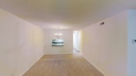 This single-floor residence offers two bedrooms and two bathrooms within a thoughtfully arranged layout. The main living and dining area is an open, combined space finished with neutral wall-to-wall carpeting that lends a warm, understated feel underfoot. A chandelier with candelabra-style arms serves as a focal point above the dining area, adding a touch of classic elegance to the otherwise clean, minimalist aesthetic. A pass-through window connects the dining area to the kitchen, providing both functional convenience and a sense of openness between the two spaces. The walls are painted in a crisp, bright white throughout, contributing to a light and airy atmosphere. A hallway visible from the main living area leads toward the bedrooms and bathrooms, suggesting a well-organized floor plan that separates private and communal spaces. The ceiling-mounted HVAC vent indicates central climate control. Consistent white trim and baseboards run throughout the space, reinforcing a cohesive and polished interior finish. Overall, the residence presents a straightforward, functional layout suited for comfortable everyday living.