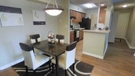 This single-floor residence offers a thoughtfully designed open-concept layout that seamlessly connects the dining and kitchen areas, creating an inviting and functional living environment. The dining area is anchored by a dark-toned table with cushioned seating for four, accented by a statement pendant light fixture overhead and a decorative area rug beneath. Warm neutral walls and hardwood-style flooring flow throughout the space, contributing to a cohesive and welcoming ambiance.

The kitchen is well-equipped with rich medium-toned cabinetry, black appliances including a refrigerator, range, and overhead microwave, and light-colored countertops that provide ample workspace. A breakfast bar peninsula separates the kitchen from the dining area, offering additional casual seating and enhancing the open feel of the shared space. Recessed and flush-mount ceiling lighting ensures the kitchen remains bright and functional.

The layout is efficient and practical, with clear sightlines between the kitchen and dining areas that make the space ideal for both everyday living and entertaining. The combination of warm wood tones, neutral walls, and modern fixtures gives the residence a balanced and comfortable aesthetic throughout.