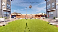 Located at 1401 Aschinger Blvd in Columbus, Ohio, this expansive single-level outdoor venue offers a generously sized open-air gathering space designed for both casual and social use. The centerpiece of the space is a large, vibrant artificial turf lawn that provides a clean, low-maintenance green surface ideal for lawn games and open-air activities — cornhole boards are visibly set up along the perimeter, inviting recreational play.

Surrounding the turf, a wood deck perimeter provides a defined gathering area furnished with multiple picnic-style tables and seating arrangements, well-suited for group dining and socializing. Large cantilever shade umbrellas are strategically positioned on both sides of the space, offering ample protection from the sun and enhancing guest comfort.

The venue is enclosed by warm-toned wooden privacy fencing, creating a sense of intimacy while maintaining a welcoming atmosphere. Overhead, a network of Edison-style string lights is strung across the open sky, lending the space a festive, inviting ambiance that transitions seamlessly from daytime to evening gatherings. A decorative pendant fixture adds an artistic focal point above the central lawn area.

The open-sky design maximizes natural light throughout the day, and the overall layout is well-organized and versatile, making this space well-suited for events, entertainment, or community use.