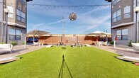 Located at 1401 Aschinger Blvd in Columbus, Ohio, this expansive single-level outdoor venue offers a generously sized open-air gathering space designed for both casual and social use. The centerpiece of the space is a large, vibrant artificial turf lawn that provides a clean, low-maintenance green surface ideal for lawn games and open-air activities — cornhole boards are visibly set up along the perimeter, inviting recreational play.

Surrounding the turf, a wood deck perimeter provides a defined gathering area furnished with multiple picnic-style tables and seating arrangements, well-suited for group dining and socializing. Large cantilever shade umbrellas are strategically positioned on both sides of the space, offering ample protection from the sun and enhancing guest comfort.

The venue is enclosed by warm-toned wooden privacy fencing, creating a sense of intimacy while maintaining a welcoming atmosphere. Overhead, a network of Edison-style string lights is strung across the open sky, lending the space a festive, inviting ambiance that transitions seamlessly from daytime to evening gatherings. A decorative pendant fixture adds an artistic focal point above the central lawn area.

The open-sky design maximizes natural light throughout the day, and the overall layout is well-organized and versatile, making this space well-suited for events, entertainment, or community use.