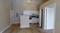 This single-floor residential space offers a straightforward, functional layout designed for comfortable everyday living. The open-concept design connects the living area to the kitchen, creating a sense of openness and ease of movement throughout the unit.

The kitchen is equipped with a white electric range and a built-in overhead range hood, complemented by an abundance of white upper cabinetry that provides generous storage space. A half-wall partition separates the kitchen from the adjacent living area, adding subtle architectural definition while maintaining the open feel. Dark countertops provide a mild contrast to the white cabinetry. An electrical panel is conveniently mounted on the wall near the kitchen partition.

The flooring transitions from tile in the kitchen area to wall-to-wall carpet in the living space, offering both practicality and comfort underfoot. The walls are finished in a neutral warm beige tone, contributing to a clean and welcoming ambiance. Ceiling-mounted track lighting illuminates the kitchen area effectively.

A doorway visible from the main living area leads to additional rooms, suggesting further private spaces within the unit. The overall layout is efficient and well-organized, making it a practical living environment.