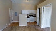 This single-floor residential space offers a straightforward, functional layout designed for comfortable everyday living. The open-concept design connects the living area to the kitchen, creating a sense of openness and ease of movement throughout the unit.

The kitchen is equipped with a white electric range and a built-in overhead range hood, complemented by an abundance of white upper cabinetry that provides generous storage space. A half-wall partition separates the kitchen from the adjacent living area, adding subtle architectural definition while maintaining the open feel. Dark countertops provide a mild contrast to the white cabinetry. An electrical panel is conveniently mounted on the wall near the kitchen partition.

The flooring transitions from tile in the kitchen area to wall-to-wall carpet in the living space, offering both practicality and comfort underfoot. The walls are finished in a neutral warm beige tone, contributing to a clean and welcoming ambiance. Ceiling-mounted track lighting illuminates the kitchen area effectively.

A doorway visible from the main living area leads to additional rooms, suggesting further private spaces within the unit. The overall layout is efficient and well-organized, making it a practical living environment.