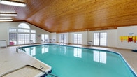 This indoor Swimming Pool and Fitness Center is a well-appointed recreational facility designed for both relaxation and active use. The pool area is the centerpiece of the space, featuring crystal-clear turquoise water set within a generously sized rectangular pool with clearly marked depth indicators for swimmer safety. A pool entry ladder provides easy access, and poolside lounge chairs are positioned along the perimeter for resting between swims. The architecture of the room is defined by a striking vaulted ceiling clad in warm natural wood planks, which lends the space a distinctive and inviting character. The ceiling's cathedral-style pitch adds to the sense of openness and volume within the room. Large grid-paned windows line the walls on multiple sides, flooding the interior with abundant natural light while offering views to the outdoors. The clean white walls complement the bright, airy atmosphere created by the combination of natural illumination and reflective pool water. The tiled pool surround provides a safe, slip-resistant walking surface around the water's edge. Overall, the space strikes a balance between functional design and aesthetic warmth, offering a year-round aquatic environment that feels both expansive and welcoming.