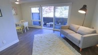 This single-floor space offers a well-designed open layout that seamlessly connects the living and dining areas. The living area features rich, dark hardwood flooring that extends throughout, complemented by a plush cream shag area rug that adds warmth and texture underfoot. A neutral-toned sofa anchors the sitting area, creating a comfortable and inviting atmosphere.

A prominent sliding glass door and wide flanking window panels flood the interior with natural light while providing direct access to a private outdoor patio. The patio is partially enclosed by fencing and features outdoor seating, making it a functional extension of the interior living space. Views of surrounding trees, including mature deciduous foliage, enhance the sense of a tranquil, nature-connected environment.

The dining area is positioned adjacent to the living space, accommodating a compact dining table and chairs, making efficient use of the open floor plan. The walls are finished in a soft, neutral tone that enhances the brightness of the space. Overall, the layout is practical and cohesive, with an airy, relaxed ambiance supported by ample natural light and a connection to the outdoors.