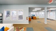 Located at 200 Berryhill Rd in Columbia, South Carolina, this single-floor commercial space offers a versatile and well-appointed layout designed to support a variety of professional uses. The interior is finished with soft blue-gray walls throughout, creating a calm and cohesive atmosphere, while neutral carpet tile flooring with teal accent squares adds a modern, functional touch underfoot.

The space is organized into distinct functional zones. A welcoming lounge area features a white sectional sofa adorned with bold navy and orange accent pillows, a vibrant area rug, and a wall-mounted flat-screen TV above a console table — ideal for client reception or informal meetings. Adjacent to this is an open work area equipped with a dedicated computer workstation and comfortable seating, bathed in natural light from multiple windows with white blinds.

A larger open workspace accommodates a full office desk setup and is surrounded by windows on multiple sides, providing abundant natural light and views of the surrounding greenery. French-style glass-paned interior doors and a structural column add architectural character, while flush-mounted fluorescent lighting ensures consistent illumination throughout. The space also includes additional rooms accessible via white panel doors, offering flexibility for private offices or storage. Overall, the layout balances collaborative and focused work environments seamlessly.