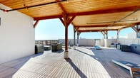 This rooftop terrace is a generously proportioned open-air amenity space defined by a striking timber-frame pergola structure. The pergola's warm, natural wood ceiling and bold exposed beams create an architectural focal point while providing partial shade across a large portion of the deck. The flooring combines light gray composite deck tiles with rich wood-tone planking near the entry, adding visual depth and texture throughout the space.

The terrace is thoughtfully arranged into distinct functional zones. A lounge area features wicker and rattan seating, offering a comfortable setting for gathering beneath the pergola. Adjacent to this is a dedicated sunbathing area with chaise lounges positioned to take full advantage of the open sky and natural light. An outdoor kitchenette with dark cabinetry, countertops, and a sink provides practical amenities for hosting, while a cornhole board adds a recreational element to the space.

A low perimeter wall with metal railing surrounds the terrace, framing sweeping panoramic views of the surrounding skyline and landscape. Access to the rooftop is provided through a set of French doors set into a crisp white exterior wall, with wall-mounted lighting flanking the entry for evening use. The combination of functional amenities, durable materials, and open sightlines makes this rooftop terrace a versatile and well-appointed outdoor space.