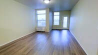 This single-floor unit features a welcoming open layout with warm hardwood-style flooring that runs throughout the main living area, lending a clean and cohesive aesthetic. The space is bathed in natural light, thanks to two generously sized windows fitted with horizontal blinds, allowing for adjustable brightness and privacy. A private entry door with a glass panel further enhances the sense of openness while providing direct access to the outdoors. The walls are finished in a neutral, light tone that complements the flooring and contributes to an airy, well-lit atmosphere. A wall-mounted PTAC heating and cooling unit is conveniently positioned beneath one of the windows, ensuring year-round climate comfort. The overall layout is straightforward and practical, offering an open canvas well-suited for a variety of furnishing arrangements. Smooth, clean finishes throughout reflect a move-in-ready condition, making this a functional and comfortable space.