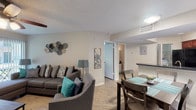 This single-floor residence offers a thoughtfully designed open-concept layout connecting the living, dining, and kitchen areas. The living room is anchored by a large sectional sofa accented with teal and patterned throw pillows, complemented by a ceiling fan with light fixture and a wall-mounted television. Vertical blinds frame a sliding glass door that opens to a private outdoor patio, allowing natural light to fill the space. Light wood-style flooring runs throughout, enhancing the bright, neutral color palette.

The kitchen is well-equipped, featuring rich cherry wood cabinetry, dark granite-style countertops, and black appliances including a refrigerator, electric range, and microwave. A double-basin stainless steel sink and a pass-through counter connecting to the dining area add both function and flow to the space.

The bedroom is generously sized, featuring direct access to the outdoor patio through a sliding glass door with vertical blinds, a walk-in closet, and hardwood-style flooring. The textured ceiling adds architectural detail consistent throughout the unit.

The bathroom includes a tub-shower combination with tile surround, a dark granite vanity countertop, and wood-style flooring, with a convenient opening adjacent to the kitchen area.