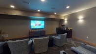Located in Woodbury, MN, this single-floor private screening room is thoughtfully designed for an immersive entertainment experience. The space centers around a large flat-screen display mounted on a neutral-toned accent wall, flanked by wall-mounted sconce lighting that casts a warm, ambient glow throughout the room. Multiple plush, upholstered lounge chairs with built-in cup holders are arranged in a semi-theater layout, providing comfortable seating for a group of viewers. The herringbone-patterned carpet adds a refined visual texture underfoot, complementing the room's sophisticated, muted color palette of grays and warm neutrals with wood-toned base trim accents. Along one wall, a built-in wet bar area features dark cabinetry with a granite countertop, a sink, and space for beverage appliances — making it convenient to enjoy refreshments without leaving the room. Decorative framed artwork adorns the side walls, contributing to the curated, lounge-like ambiance. Recessed ceiling lighting works in concert with the sconces to allow flexible mood control. The overall design strikes a balance between the comfort of a living room and the functionality of a dedicated screening space, making it ideal for movie nights or private viewings.