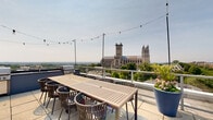 Located at 3201 Wisconsin Ave NW in Washington, DC, this expansive rooftop terrace offers an elevated outdoor retreat with sweeping panoramic views of the city skyline and surrounding neighborhoods. The terrace is finished with large-format pavers throughout, lending a clean and contemporary aesthetic to the open-air space.

The rooftop is thoughtfully divided into distinct functional zones. A comfortable lounge area features plush blue sectional seating and chaise lounges, ideal for relaxing while taking in the broad cityscape. Nearby, a dedicated outdoor dining area accommodates a large rectangular table with woven rattan chairs, perfect for al fresco gatherings. String lights are strung overhead, adding a warm ambiance for evening use.

A well-equipped grilling station lines one side of the terrace, offering multiple gas grills for outdoor cooking. Lush plantings in oversized blue ceramic pots are strategically placed throughout, introducing vibrant seasonal color and greenery. Low-profile planters along the perimeter railing further enhance the garden-like atmosphere.

The glass and metal railing system ensures unobstructed sightlines across Washington's treeline and mid-rise urban fabric. The building's dark brick mechanical structure anchors the rooftop's northwest corner, adding architectural character. This rooftop terrace serves as a premier outdoor amenity space in the heart of the Cleveland Park neighborhood.