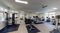 This fitness center is a thoughtfully designed, single-floor facility offering a comprehensive workout environment across multiple open areas. The space features light wood-style flooring throughout, crown molding, and a neutral color palette of soft whites and warm grays that create a clean, inviting atmosphere.

The main workout area is generously proportioned and lined with floor-to-ceiling mirrors that amplify natural light and enhance the sense of space. Multiple treadmills, elliptical machines, and stationary bikes are arranged across rubber floor mats, complemented by a multi-function cable machine and a rack of dumbbells for strength training. Ceiling fans provide additional air circulation throughout the workout zones.

A separate functional training area includes a wall-mounted flat-screen TV, stability balls, medicine balls, resistance bands, and a cable strength machine — ideal for versatile, full-body workouts. An additional room houses a stationary spin bike and offers views into adjacent workout areas through interior windows, with natural light streaming in from multiple exterior windows.

The facility also includes a dedicated restroom, finished with large-format white tile flooring, a pedestal sink, a round mirror, and a bold teal accent wall accented by decorative open shelving — adding a touch of style to the functional space. A water dispenser is also available on the fitness floor for convenience.