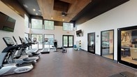 Located at 917 7th Avenue in Garner, North Carolina, this single-floor amenity facility offers a thoughtfully designed space combining fitness and productivity. The fitness center is the centerpiece of the space, boasting soaring ceilings with a striking wood-paneled soffit detail and recessed lighting. Large clerestory windows along the upper walls flood the gym with natural light and offer views of the surrounding greenery, creating an energizing workout atmosphere. The rubber-floored gym is equipped with a full array of cardio machines, including treadmills, ellipticals, and a rowing machine, as well as a dedicated free weights area featuring a multi-tier dumbbell rack and adjustable bench. Wall-mounted flat-screen televisions and a ceiling fan add to the comfort of the workout space. Black-framed glass doors connect zones within the facility seamlessly. Adjacent to the gym is a vibrant co-working and business center, distinguished by bold orange walls, a grid-pattern accent partition, and light wood-look flooring. This productive nook features computer workstations, collaborative seating around a round table, and a pendant light, making it well-suited for both focused work and casual meetings. Together, these spaces offer a dynamic and modern environment for fitness and work.