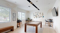 This single-floor work room offers a well-appointed and versatile space designed for both recreation and casual gatherings. The layout is anchored by a central game room featuring a full-size pool table with a rich wood frame and neutral felt surface, accompanied by a dedicated cue rack and a wall-mounted flat-screen television — ideal for entertaining or unwinding.

The interior design strikes a clean, modern aesthetic with crisp white walls adorned with elegant tropical palm leaf murals in soft gold tones. A curated gallery wall of framed artwork and decorative accents adds personality and warmth to the space. Large windows fitted with horizontal blinds line the walls, flooding the room with abundant natural light and offering views of the surrounding greenery outdoors.

Large-format light-colored tile flooring runs throughout, lending a seamless and open feel. Seating options include a wooden bench along the perimeter and a bistro-style high-top table with bar chairs, creating an inviting nook for conversation or casual work. Recessed ceiling lighting alongside a sleek track light fixture ensures well-distributed illumination across the room.

A connected bedroom and a corridor with secure dark-framed glass entry doors round out this thoughtfully designed space, which balances functional amenities with a welcoming, resort-inspired ambiance.