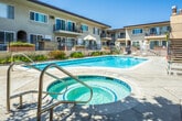 Located at 20455 Anza Ave in Torrance, California, this outdoor communal area serves as the centerpiece of a multi-building residential complex. The space is anchored by a generously sized swimming pool with clear, blue water, complemented by a separate circular spa/hot tub positioned nearby. The pool deck is finished with light-toned concrete paving and is outfitted with multiple lounge chairs and patio umbrellas, providing ample space for sunbathing and relaxation under the Southern California sun.

The surrounding buildings feature a stucco exterior in a warm beige tone with accents of Spanish-influenced architecture, including arched entryways, tile roofing details, and wrought iron balcony railings. Several units feature private balconies that overlook the pool area. Exterior staircases with metal railings provide access between floors of the complex. Lush landscaping with shrubs, flowering plants, and mature trees borders the pool enclosure, adding greenery and privacy to the outdoor setting.

The pool area is enclosed by a metal security fence, with a dedicated utility room and signage indicating pool safety guidelines. The overall ambiance is bright, open, and inviting, characteristic of Southern California outdoor living at its finest.
