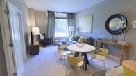 This single-floor residence offers an open-concept layout that thoughtfully integrates living and dining areas into one cohesive space. The living area features a plush blue-grey sofa accented with an assortment of decorative pillows in blue, green, and gold tones, complemented by a deep navy wingback chair. A large framed artwork in muted gold tones anchors the wall, adding a sophisticated focal point. Neutral grey walls and plush carpeted flooring contribute to a calm, cohesive aesthetic throughout. A generous window dressed with elegant floor-length grey curtains allows natural light to filter in, offering views of lush greenery outside. The dining area features a round white-topped table with a distinctive sculptural wood base, surrounded by modern chartreuse chairs that introduce a vibrant yet tasteful pop of color. Layered area rugs define each zone while maintaining visual continuity. Thoughtful lighting — including a tall white floor lamp and a matching table lamp — ensures warm, ambient illumination throughout. Overall, the space strikes a balance between contemporary design and relaxed comfort.
