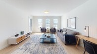 This well-appointed single-floor residence offers a thoughtfully designed open-concept layout that seamlessly connects the kitchen, dining, and living areas. The kitchen is a standout feature, equipped with white shaker cabinetry, granite countertops, a large island with bar seating, stainless steel appliances including a refrigerator, range, microwave, and dishwasher, and elegant globe pendant lighting. Light hardwood flooring runs throughout the main living areas, enhancing the bright, airy atmosphere created by multiple windows that invite generous natural light. The living area provides ample room for comfortable seating arrangements, while the adjacent dining space accommodates a full dining set. Two carpeted bedrooms each feature multiple windows with exterior views, offering a calm and restful environment. The full bathroom includes large-format tile flooring, a walk-in shower, and clean white finishes throughout. A dedicated walk-in closet provides extensive storage with built-in shelving and double hanging rods. Adding to the unit's practicality, an in-unit stacked washer and dryer is tucked neatly behind a closet door. The overall design maintains a clean, contemporary aesthetic with a neutral white palette and modern fixtures throughout.