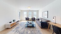 This well-appointed single-floor residence offers a thoughtfully designed open-concept layout that seamlessly connects the kitchen, dining, and living areas. The kitchen is a standout feature, equipped with white shaker cabinetry, granite countertops, a large island with bar seating, stainless steel appliances including a refrigerator, range, microwave, and dishwasher, and elegant globe pendant lighting. Light hardwood flooring runs throughout the main living areas, enhancing the bright, airy atmosphere created by multiple windows that invite generous natural light. The living area provides ample room for comfortable seating arrangements, while the adjacent dining space accommodates a full dining set. Two carpeted bedrooms each feature multiple windows with exterior views, offering a calm and restful environment. The full bathroom includes large-format tile flooring, a walk-in shower, and clean white finishes throughout. A dedicated walk-in closet provides extensive storage with built-in shelving and double hanging rods. Adding to the unit's practicality, an in-unit stacked washer and dryer is tucked neatly behind a closet door. The overall design maintains a clean, contemporary aesthetic with a neutral white palette and modern fixtures throughout.