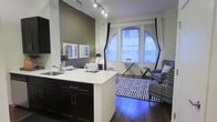 This thoughtfully designed single-floor residence showcases a seamless open-concept layout that connects the kitchen and living areas into one cohesive, functional space. The kitchen features dark espresso cabinetry with sleek hardware, a crisp white countertop island with an integrated sink, and track lighting overhead that provides focused illumination across the workspace. A bold dark accent wall anchors the space and adds depth, complemented by a pair of framed artwork pieces that bring a curated aesthetic to the interior. The living area is anchored by a striking geometric-patterned area rug in black and white, which plays well against the rich hardwood flooring beneath. A patterned accent chair and a minimalist side table contribute to the room's contemporary character. The standout architectural feature is the dramatic arched window, which floods the living space with abundant natural light and serves as a focal point of the room. White curtains frame the window elegantly. Track lighting and ambient accent lighting throughout add layers of warmth and functionality. The overall ambiance is modern and urban, with a confident design palette of dark tones, crisp whites, and bold patterns that come together to create a polished and inviting living environment.