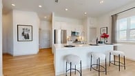 This well-appointed single-floor residence offers a thoughtfully designed open-concept layout that maximizes both function and comfort. The kitchen is a standout feature, showcasing white shaker cabinetry reaching to the ceiling, a classic subway tile backsplash, stainless steel appliances including a French door refrigerator, over-range microwave, and range, all anchored by a generous island with quartz countertops and bar seating — ideal for casual dining or entertaining. Light wood-tone flooring flows throughout the main living areas, enhancing the bright, airy atmosphere created by recessed lighting and an abundance of natural light. The living room is generously scaled, centered around large sliding glass doors that open to a private balcony with open outdoor views. A built-in mudroom/cubby area near the entry provides practical storage with cubbies, hooks, and shelving. The bedroom features plush carpeting, a ceiling fan, and a large window with natural light. The bathroom is finished with white cabinetry, quartz counters, and a tiled tub-shower combination. Clean, contemporary design and neutral tones throughout give the space a cohesive, modern feel.