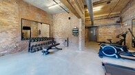 Located at 250 S Depot St in Pendleton, South Carolina, this fitness center combines industrial character with functional design. The space is defined by its striking exposed brick walls, which lend a raw, historic aesthetic throughout the room. Soaring ceilings with exposed ductwork and conduit add to the industrial ambiance, while polished concrete floors provide a durable, clean foundation underfoot.

Natural light pours in through large double-door windows along one wall, illuminating the space and offering views of the surrounding greenery. The layout is thoughtfully organized to accommodate a variety of workout modalities. Cardio equipment — including treadmills, stationary bikes, and rowing machines — is arranged along the perimeter, complemented by wall-mounted flat-screen televisions. A dedicated free weights area features a full rack of dumbbells and kettlebells, an adjustable bench, and a wall mirror for form checking. Yoga mat storage and additional fitness accessories are neatly mounted to the brick walls.

Large framed mirrors are strategically placed to enhance the sense of space and support training. The combination of exposed masonry, high ceilings, and abundant natural light creates an energizing yet characterful environment well-suited for a comprehensive fitness routine.