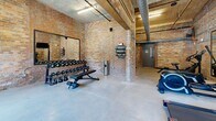 Located at 250 S Depot St in Pendleton, South Carolina, this fitness center combines industrial character with functional design. The space is defined by its striking exposed brick walls, which lend a raw, historic aesthetic throughout the room. Soaring ceilings with exposed ductwork and conduit add to the industrial ambiance, while polished concrete floors provide a durable, clean foundation underfoot.

Natural light pours in through large double-door windows along one wall, illuminating the space and offering views of the surrounding greenery. The layout is thoughtfully organized to accommodate a variety of workout modalities. Cardio equipment — including treadmills, stationary bikes, and rowing machines — is arranged along the perimeter, complemented by wall-mounted flat-screen televisions. A dedicated free weights area features a full rack of dumbbells and kettlebells, an adjustable bench, and a wall mirror for form checking. Yoga mat storage and additional fitness accessories are neatly mounted to the brick walls.

Large framed mirrors are strategically placed to enhance the sense of space and support training. The combination of exposed masonry, high ceilings, and abundant natural light creates an energizing yet characterful environment well-suited for a comprehensive fitness routine.