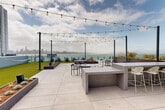 Located on Treasure Island in San Francisco, this rooftop terrace delivers a striking outdoor experience defined by sweeping, unobstructed views of the San Francisco Bay, the city skyline, and the Bay Bridge. The space is finished with large-format porcelain pavers throughout, lending a clean, contemporary aesthetic that ties the various zones together seamlessly.

The terrace is thoughtfully organized into multiple functional areas. A turf lawn provides a casual relaxation zone, outfitted with bean bag seating and bordered by raised planter beds filled with native and ornamental plantings. Overhead string lights crisscross the main gathering area, adding ambient character. Concrete communal tables and chair seating clusters offer spaces for both dining and socializing, while a dedicated lounge area features upholstered outdoor furniture ideal for quieter settings.

A glass railing system runs along the perimeter, preserving sightlines and maximizing the visual connection to the bay. Decorative bronze sculptural elements mounted on a perimeter wall add an artistic dimension to the space. Additional planter beds line a quieter wing of the terrace, which leads to an indoor access structure with glass doors. The overall design balances open gathering space with intimate pockets, making this rooftop a highly functional and visually remarkable outdoor destination.