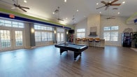 This vibrant single-floor amenity lounge is designed for socializing and recreation, offering an open, airy layout defined by soaring vaulted ceilings and multiple ceiling fans that enhance the sense of volume and comfort. The main area is anchored by a full-size pool table centered on smooth hardwood-style flooring, providing ample space for leisure and entertainment. Bold navy and lime green accent bands run along the upper walls, giving the space a lively, energetic character.

Large windows with plantation shutters and multiple sets of French doors line the walls, flooding the interior with natural light and offering views of the surrounding outdoor areas. Wall-mounted sconces complement the recessed lighting throughout for a well-lit atmosphere at any time of day.

A bar-height counter with wood-toned seating provides a casual gathering spot, while a wall-mounted flat-screen TV and a Coca-Cola vending machine add convenient amenities for guests. Decorative indoor plants and a patterned accent wall with a package locker system add functional and aesthetic interest.

Adjacent to the main lounge is a fully equipped kitchen featuring dark wood cabinetry, light stone countertops, stainless steel appliances including a refrigerator, range, and dishwasher, and tile flooring — ideal for catering or community events. The entry foyer transitions seamlessly from tile to hardwood flooring, setting a welcoming tone from the moment of arrival.