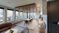This thoughtfully designed single-floor space showcases a seamless open-concept layout that elegantly connects a fully equipped kitchen, a generous dining area, and a relaxed lounge zone. A defining architectural feature is the slatted wood ceiling treatment that spans the dining and living areas, adding warmth and visual texture to the contemporary interior. Pendant lights with dark glass shades hang above a solid wood dining table surrounded by plush green velvet chairs, creating a refined yet inviting gathering space. Large windows wrap the perimeter, flooding the interior with natural light and offering expansive views of the surrounding urban landscape. The lounge area features a built-in fireplace set within a concrete-style surround, complemented by a low wooden media shelf and an eclectic collection of organic wall art pieces. The kitchen is a standout, boasting a bold dark backsplash tile, open floating shelves, matte grey cabinetry, stainless steel appliances, and a substantial island with a richly veined stone countertop and gas cooktop. Light hardwood flooring flows throughout, tying the spaces together cohesively. The overall ambiance is modern and sophisticated, balancing natural materials with sleek finishes for a polished, livable aesthetic.