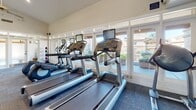 Located on S Bill Martin Dr in Tucson, AZ, this single-floor fitness center offers a well-equipped space designed to support a comprehensive workout experience. The facility features vaulted ceilings fitted with ceiling fans and recessed lighting, contributing to an open, airy atmosphere throughout the main exercise area.

The cardio zone is positioned along a wall of large windows and glass doors, allowing abundant natural light and views of the surrounding outdoor landscape. This section houses multiple treadmills and elliptical machines, each equipped with individual monitors. The strength training area includes a dumbbell rack with a full range of free weights, an adjustable workout bench, and cable machines, all positioned in front of floor-to-ceiling mirrors that enhance the sense of space.

Additional equipment includes stationary bikes and a functional training station. A dedicated stretch and accessory area near the entrance features a tiered stability ball rack holding multiple sized balls. The facility is accessed through double glass doors marked with fitness center signage, and the interior is finished with durable carpet tile flooring throughout. The overall design balances functionality with a clean, bright aesthetic suited for daily use.