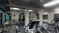 This fitness center offers a comprehensive workout environment designed to accommodate a wide range of exercise routines. The space is organized across a single floor and is clearly divided into distinct training zones, making it easy to navigate between cardio and strength-focused areas.

The cardio section is outfitted with multiple machines, including treadmills, ellipticals, and a stationary cycle, providing ample options for aerobic training. Adjacent to this area, a dedicated strength training zone houses a multi-station cable machine, a punching bag, and a selection of free weights and dumbbells neatly arranged on a rack — ideal for both resistance and functional training.

Large mirrored walls line portions of the space, enhancing the sense of openness while allowing users to monitor their form during workouts. Structural columns are integrated throughout the floor plan, adding an architectural element that subtly segments the space without interrupting the overall flow.

Overhead fluorescent lighting ensures the entire area is well-lit, contributing to an energetic and focused atmosphere. The flooring features a dark, rubberized surface suited for high-impact activities. Teal accent walls add a pop of color, giving the facility a clean, modern aesthetic. Informational workout charts are posted on the walls as a practical resource for users.