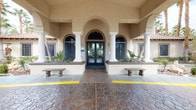 This single-story senior living community facility presents an inviting and thoughtfully designed environment from the moment of arrival. The exterior entrance showcases a Mediterranean-inspired architectural style, featuring stucco arches, white columns, arched dark-framed windows, and decorative stonework paving, flanked by stone benches and tropical landscaping that set a warm, resort-like tone.

Inside, the central lobby serves as the heart of the facility. Soaring vaulted ceilings draw the eye upward to a striking double-ring chandelier with cascading crystal elements, which anchors the space with elegance. A dramatic floor-to-ceiling dark accent fireplace wall serves as a focal point, while comfortable wingback chairs arranged around a round wooden table encourage relaxed conversation. Crown molding, recessed lighting, and light wood-look flooring unify the aesthetic throughout. A "Wall of Honor" display is visible to one side, suggesting a space that celebrates residents' histories and community pride.

The adjacent dining and common area is generously proportioned, featuring coffered ceilings with ceiling fans, multiple dining tables and chairs, and an open-plan kitchen equipped with modern appliances. Arched doorways connect the spaces fluidly, and natural light filters in through windows, contributing to an open, airy ambiance well-suited to communal dining and daily activities.