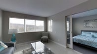 This single-floor residence offers a thoughtfully laid-out living space with a clean, contemporary aesthetic throughout. The living area is a standout feature, bathed in natural light streaming through a wide bank of windows fitted with horizontal blinds. Neutral gray walls, light-toned flooring, and smooth ceilings create a calm, cohesive atmosphere. A glass-top coffee table and area rug anchor the seating area, while a chevron-patterned accent chair adds a subtle decorative touch near the windows.

A sliding glass-panel door connects the living area to the bedroom, which features darker flooring and a decorative headboard, offering a private retreat while maintaining a visual connection to the main living space. The open sightlines between rooms contribute to a sense of spaciousness throughout.

The kitchen area, positioned toward the rear of the floor plan, is outfitted with white cabinetry and appears well-organized, providing practical functionality. The overall layout flows naturally from the living and dining zones through to the sleeping and cooking areas, making efficient use of the available space. The consistent use of neutral tones and modern finishes throughout ties each area together into a unified, welcoming environment.