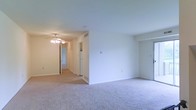 This single-floor space presents a clean, functional layout with a straightforward and practical design. The main living area is finished with wall-to-wall neutral-toned carpet, offering a soft underfoot feel that extends throughout the space. Bright white walls provide a blank canvas that allows for flexible interior styling, while standard-height ceilings keep the space feeling open and manageable.

A ceiling fan with an integrated light fixture is prominently positioned overhead, serving both as a lighting source and an aid for air circulation. Overhead HVAC vents are visible, indicating a centralized climate control system throughout the unit.

A hallway extends from the main living area, connecting to additional rooms deeper within the space. From this vantage point, a white refrigerator is visible at the end of the corridor, indicating the presence of a kitchen area. The hallway is lined with the same neutral carpeting and white walls, maintaining a consistent aesthetic throughout.

Overall, the space offers a clean, move-in-ready feel with minimal ornamentation, making it highly adaptable to various furnishing styles and functional needs.