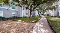 This residential community exterior presents a welcoming, well-kept environment defined by its classic architectural design and thoughtfully maintained grounds. The multi-story building features a neutral gray lap siding facade complemented by white-trimmed balconies and railings, lending a clean, cohesive aesthetic throughout. Ground-floor units offer private covered porches, while upper-level residences enjoy open balconies that overlook the landscaped courtyard below.

The grounds are a standout feature, with mature live oak trees providing generous shade across the property. Their broad canopies arch naturally over the concrete walkways that wind through the community, creating a peaceful, tree-lined corridor between buildings. Neatly manicured grass, decorative ground cover, and low-maintenance shrub beds line the building perimeters, adding layers of greenery that enhance the park-like ambiance.

Concrete pathways connect the various sections of the property, offering easy pedestrian access throughout. Decorative stone mulch beds are interspersed with plantings, contributing to the clean and organized landscaping scheme. Exterior stairwells provide access to upper floors, while the overall site layout promotes an open, airy feel with ample space between structures. The community conveys a tranquil, residential character enriched by its natural surroundings.
