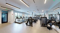 Located at 590 NW 159th St in Miami, FL, this professionally equipped fitness center spans a generous single floor, offering a comprehensive workout environment designed for a wide range of training needs. The space is defined by its clean, modern aesthetic, featuring light wood-style flooring, crisp white walls, and sleek linear pendant lighting that runs throughout the ceiling, creating a bright and energizing atmosphere. Large floor-to-ceiling windows line the perimeter, flooding the main exercise area with natural light and providing open views of the surrounding landscape. The primary workout floor is outfitted with an extensive array of commercial-grade strength machines, including multi-functional cable systems such as the Synergy 360, selectorized weight machines, free weight benches, and a full rack of dumbbells. A dedicated cardio zone features multiple treadmills and upright bikes arranged in a well-spaced row. A separate studio area, enclosed by black-framed glass partition walls, provides space for cycling classes and group training, with floor-to-ceiling mirrors amplifying the sense of openness. An additional lounge-like zone showcases a decorative wood slat accent wall with a mounted flat-screen display, adding a warm, design-forward touch to the space. Throughout, the layout ensures ample circulation space between stations, supporting a comfortable and functional fitness experience.