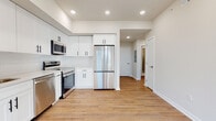 This single-floor unit showcases a clean, modern aesthetic carried consistently throughout its layout. The kitchen serves as a functional centerpiece, outfitted with white shaker-style cabinetry accented by matte black hardware, white quartz countertops, and a full suite of stainless steel appliances including a French door refrigerator, electric range, over-the-range microwave, and dishwasher. Recessed lighting and light-toned hardwood flooring complete the space, which opens toward a hallway connecting the remaining rooms.

The bedrooms are bright and neutrally finished, each featuring large double-hung windows that invite natural light and offer exterior views of surrounding greenery and urban surroundings. Hardwood flooring flows throughout these rooms, complementing the warm, minimalist palette of beige and white walls.

The bathrooms are well-appointed with distinct finishes. One bathroom features crisp white hexagonal floor tile, a white vanity, and a window providing natural light. A second bathroom includes large-format gray wall tile, a glass-enclosed walk-in shower with hexagonal mosaic tile flooring, and chrome fixtures, lending a more spa-like feel. Both spaces are clean, modern, and efficiently designed.