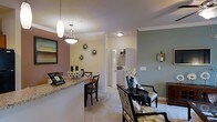 Located at 1150 Baxter Lane in Knightdale, NC, this single-floor residence offers a well-designed, open-concept living space that balances comfort and style. Crown molding throughout adds a refined architectural detail, lending the interior a polished, cohesive feel.

The living and dining areas flow seamlessly together, creating an inviting, connected atmosphere. A granite-topped breakfast bar serves as a natural divider between the kitchen and dining space, offering casual seating alongside a dedicated dining area illuminated by a stylish pendant light fixture. The soft, neutral carpet in the living area provides warmth underfoot, while the muted sage and warm beige wall tones contribute to a calm, welcoming ambiance.

Decorative wall accents, including sunburst mirrors and framed artwork, add character without overwhelming the space. The sitting area is furnished with patterned upholstered chairs and a round accent table, creating an intimate nook for relaxing or entertaining.

A glimpse into the utility area reveals an in-unit washer and dryer, a highly practical feature. Closet storage is also visible, suggesting thoughtful use of available space throughout this efficiently designed single-level home.