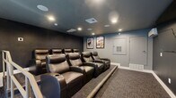 Located in Escondido, CA, this private theater delivers an authentic cinematic experience within a well-appointed, intimate setting. The space is designed with a distinct two-zone layout: a welcoming entry area and a dedicated screening room, each thoughtfully finished to enhance the movie-going atmosphere.

The entry area greets guests with dark, charcoal-toned walls, carpet flooring, and a classic red popcorn machine that sets the tone immediately upon arrival. Double glass-paneled doors with blue frames provide access, while a handrail-equipped ramp connects the entry to the main screening level. A compact refrigerator offers convenient refreshment storage, and safety features including an illuminated exit sign and fire extinguisher are prominently placed.

The screening room itself is anchored by a row of plush, dark brown leather recliner seats with built-in cup holders, arranged on a raised platform for optimal sightlines. Framed vintage-style movie posters adorn the lighter gray accent wall, complementing the deep charcoal ceiling and surrounding walls. Recessed lighting provides a warm, ambient glow throughout. In-ceiling speakers suggest a well-integrated audio system, and a wall-mounted screen completes the cinematic setup. The combination of purposeful design and quality furnishings makes this a standout entertainment space.