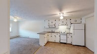 Located at 1 Morningside Ct in Salem, New Hampshire, this single-floor unit offers a straightforward and functional layout designed for comfortable everyday living. The kitchen is outfitted with white cabinetry accented by black hardware, a tile backsplash, granite-style countertops, and a full suite of white appliances including an electric range, built-in microwave, and refrigerator. Tile flooring runs throughout the kitchen area, while a peninsula counter provides a natural transition into the open living space. The living area features soft wall-to-wall carpeting, a ceiling fan with light, and a baseboard heating system. Windows along the exterior wall allow natural light to fill the room and offer views of the surrounding greenery. A dedicated bedroom continues the carpeted flooring and includes its own ceiling fan, windows with outdoor views, and baseboard heat. The full bathroom is finished with white tile surrounding the tub-shower combination, a white vanity with sink, and a medicine cabinet with mirror. Throughout the unit, the clean white walls and consistent finishes create a bright, neutral aesthetic that is easy to personalize.