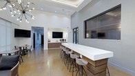 Located at 511 Cobblestone Dr in Wilmington, North Carolina, this single-floor space offers a sophisticated and contemporary design that blends functionality with modern aesthetics. The main gathering area immediately impresses with its soaring ceilings, enhanced by a recessed tray ceiling detail and flush lighting that creates a bright, open atmosphere throughout.

A standout feature of the space is the substantial bar-height counter, clad in warm wood paneling and topped with a crisp white surface, accompanied by a row of stylish industrial-style bar stools. This inviting setup is ideal for social gatherings or casual meetings. Multiple wall-mounted flat-screen TVs are strategically positioned throughout the space, ensuring excellent visibility from multiple vantage points.

The flooring consists of polished concrete in a warm earth tone, lending an industrial yet refined character to the overall design. The walls are finished in clean white and soft gray tones, contributing to the space's bright and airy feel. Built-in cabinetry in a warm wood finish along one wall provides additional storage and functionality, complemented by beverage station amenities.

A connecting hallway extends the space further, with additional rooms visible beyond, suggesting versatile, multi-use functionality. Large dark-framed windows add architectural interest while connecting the interior to the outdoors.
