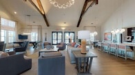 This single-floor community space is defined by its dramatic vaulted ceilings adorned with rustic exposed wood beams, creating a striking architectural contrast against the clean white walls and wide-plank hardwood flooring that flows throughout. The expansive main lounge area is designed for social gatherings, featuring multiple seating arrangements with plush sofas, accent chairs, and stylish console tables. A branching chandelier serves as a focal art piece overhead, while large black-framed French doors line the walls, flooding the interior with natural light and providing direct access to an outdoor terrace. A dedicated game area includes a shuffleboard table and a pool/foosball table, accented by bold abstract artwork on the walls. The kitchen is well-appointed with white shaker cabinetry, dark subway tile backsplash, stainless steel appliances, and a large center island with seating — ideal for catering or casual use. A cozy lounge nook features a floor-to-ceiling dark accent wall with a fireplace, framed artwork, and comfortable seating arrangements. A private dining area accommodates intimate gatherings beneath a sculptural chandelier, with patterned drapery and garden views through large windows. Adjacent glass-enclosed conference or meeting rooms provide flexible private space. The overall ambiance is warm, modern, and inviting.