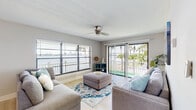 This thoughtfully designed single-floor residence offers a seamless blend of coastal charm and modern comfort. The living room serves as the heart of the space, featuring light hardwood-style flooring, a ceiling fan, and an abundance of natural light streaming through large horizontal-blind windows and sliding glass doors that open to a private balcony. From here, sweeping waterfront views and lush palm trees create a serene, resort-like atmosphere.

The dining area is elegantly appointed with a marble-top round table, modern white chairs, and a circular pendant light fixture, complemented by contemporary wall art. The galley-style kitchen is well-equipped with stainless steel appliances, crisp white shaker cabinetry, clean countertops, and light hardwood-style flooring throughout.

The primary bedroom is generously sized, showcasing a bold navy upholstered bed frame, abstract wall art, and in-unit laundry access. The second bedroom is bright and airy, with a garden window view framed by palm foliage and its own distinctive artwork. Both bathrooms are finished with marble-style tile accents, tub-shower combinations, and wood-look flooring, offering a clean, contemporary aesthetic throughout.